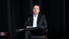 Elon Musk अमेरिकी एयर फोर्स एकेडमी के कार्यक्रम में काले सूट में कुर्सी पर बैठे हुए