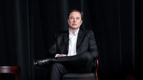 Elon Musk अमेरिकी एयर फोर्स एकेडमी के कार्यक्रम में काले सूट में कुर्सी पर बैठे हुए